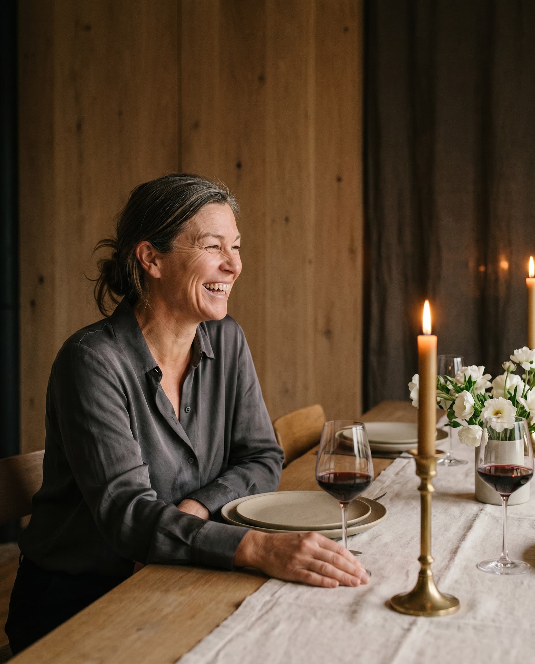The same woman is mid-laugh in conversation at a candlelit dinner, wearing a charcoal silk blouse, brass candlestick and wine glasses on the linen-set table beside her.