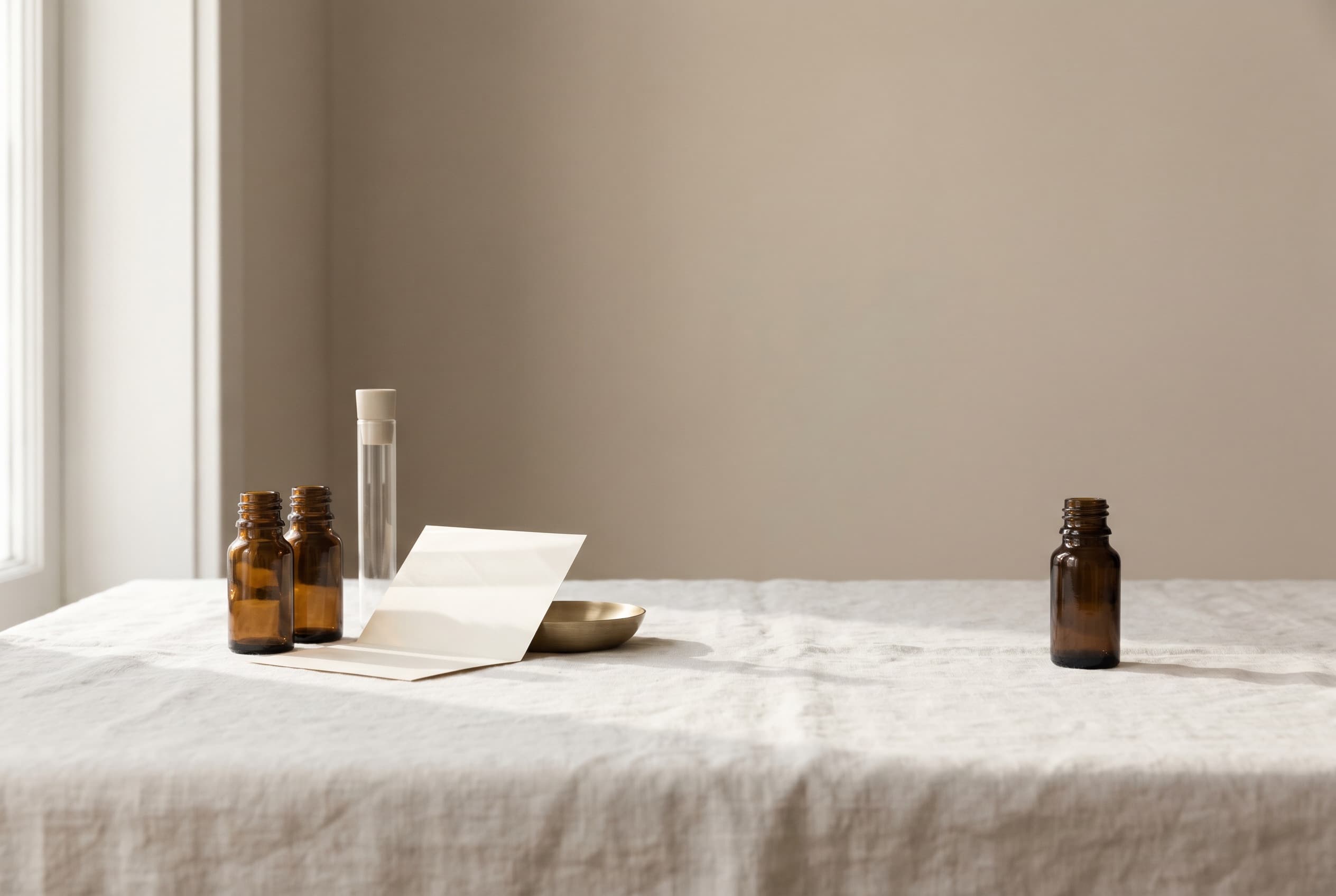 An arrangement of vials, brass dishes, and folded paper beside a single separate vial on bone linen.